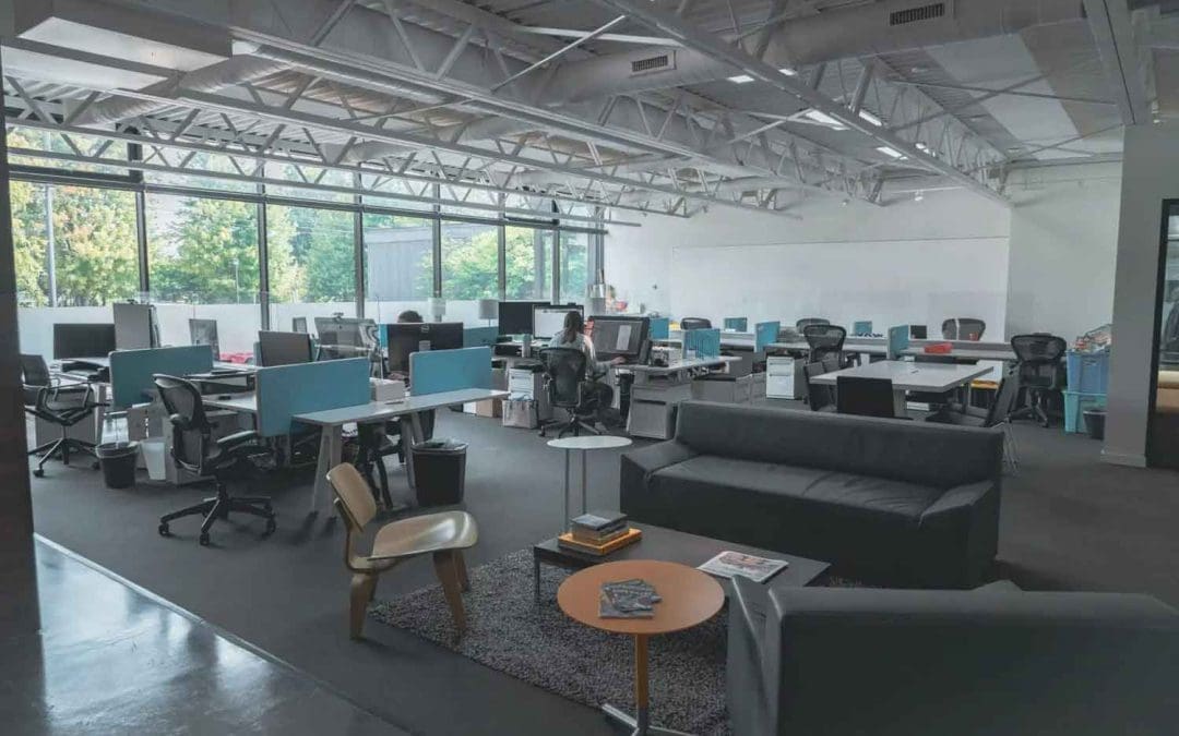 A wide view of a large, open-plan office space featuring rows of desks with computers, ergonomic chairs, and a comfortable seating area with a sofa and coffee table in the foreground.