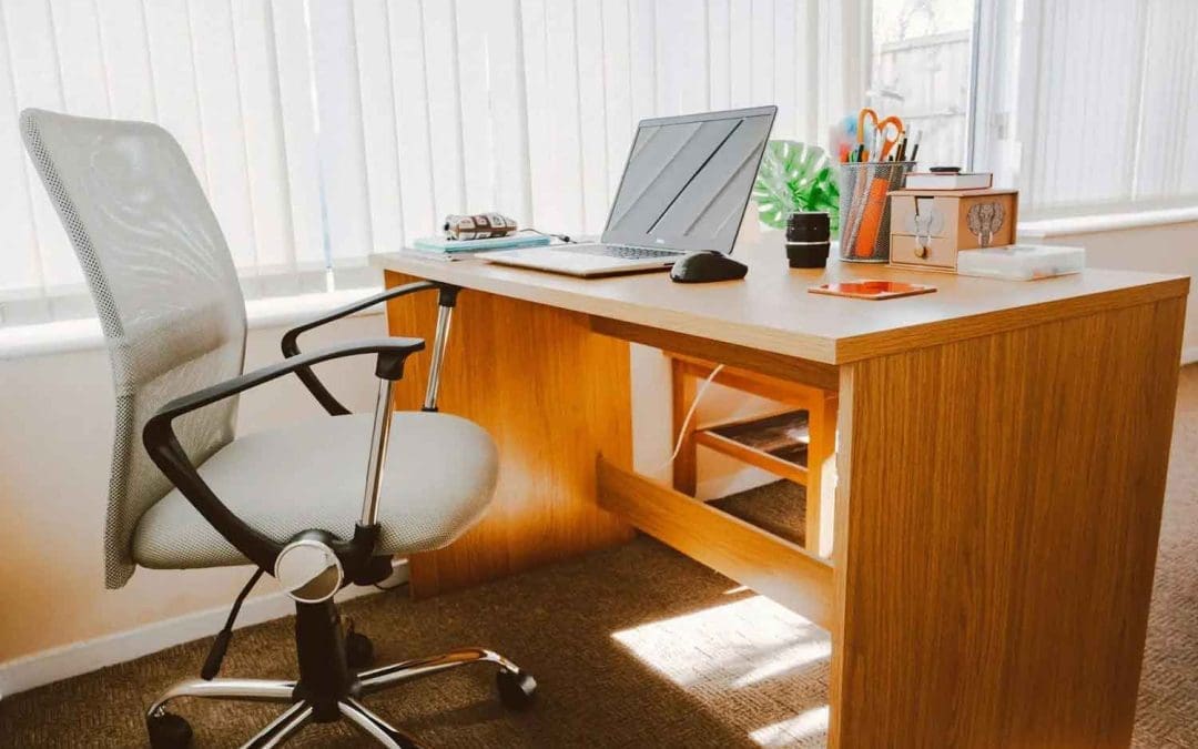 A cozy office setup with a wooden desk, ergonomic chair, laptop, mouse, and potted plant, illuminated by natural light from large windows with white blinds.