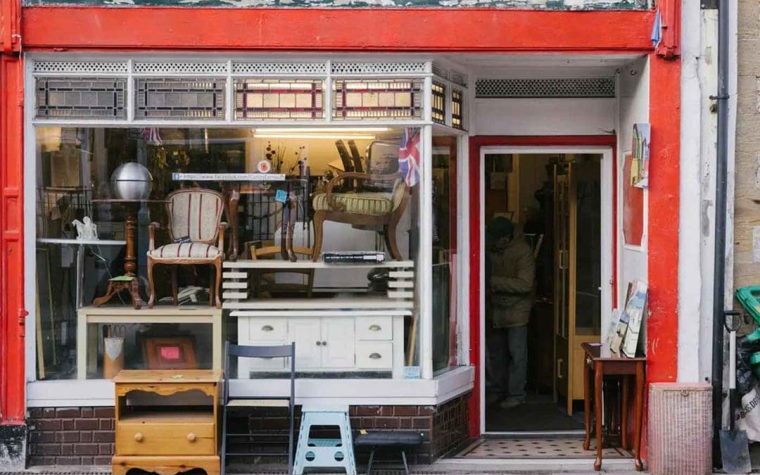 A quaint antique shop with a red frame, displaying vintage furniture like chairs and tables in the window, with a Union Jack flag and a person entering the store.